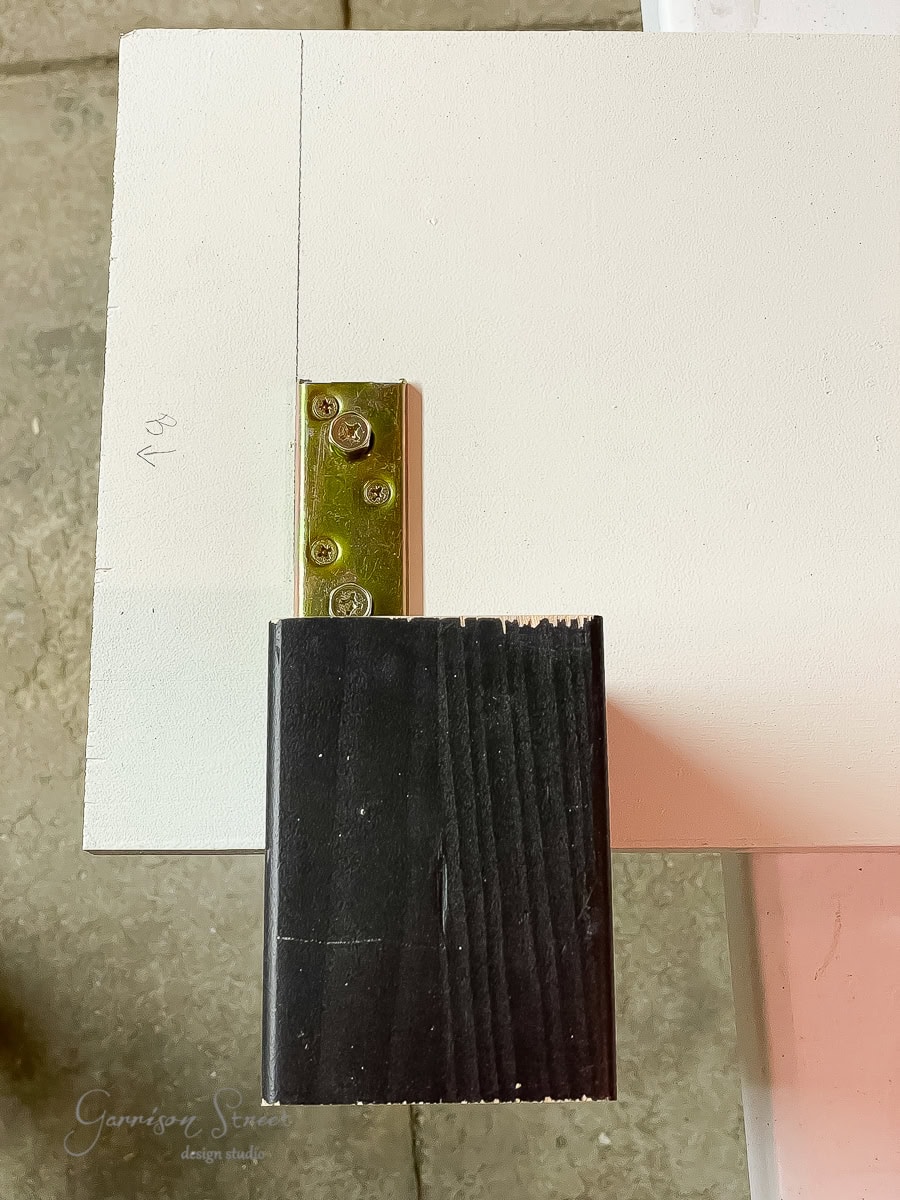 A metal hinge is attached to the edge of a white wooden board, resembling an adjustable base. A black wooden block is positioned below the board, suggesting elements of a bed frame. The setup rests on a rough, gray surface.