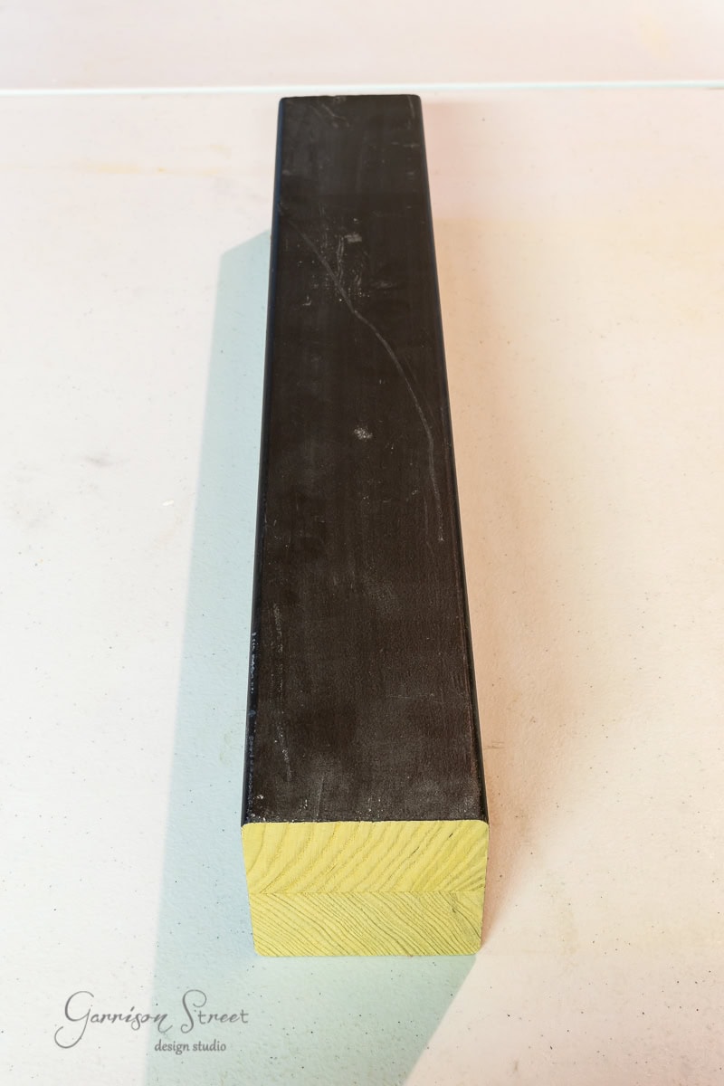 A long rectangular wooden beam with a black surface is elegantly positioned on a light-colored table, resembling an adjustable base. The grain of the wood is visible at the end, and a logo for Garrison Street Design Studio graces the bottom left corner.