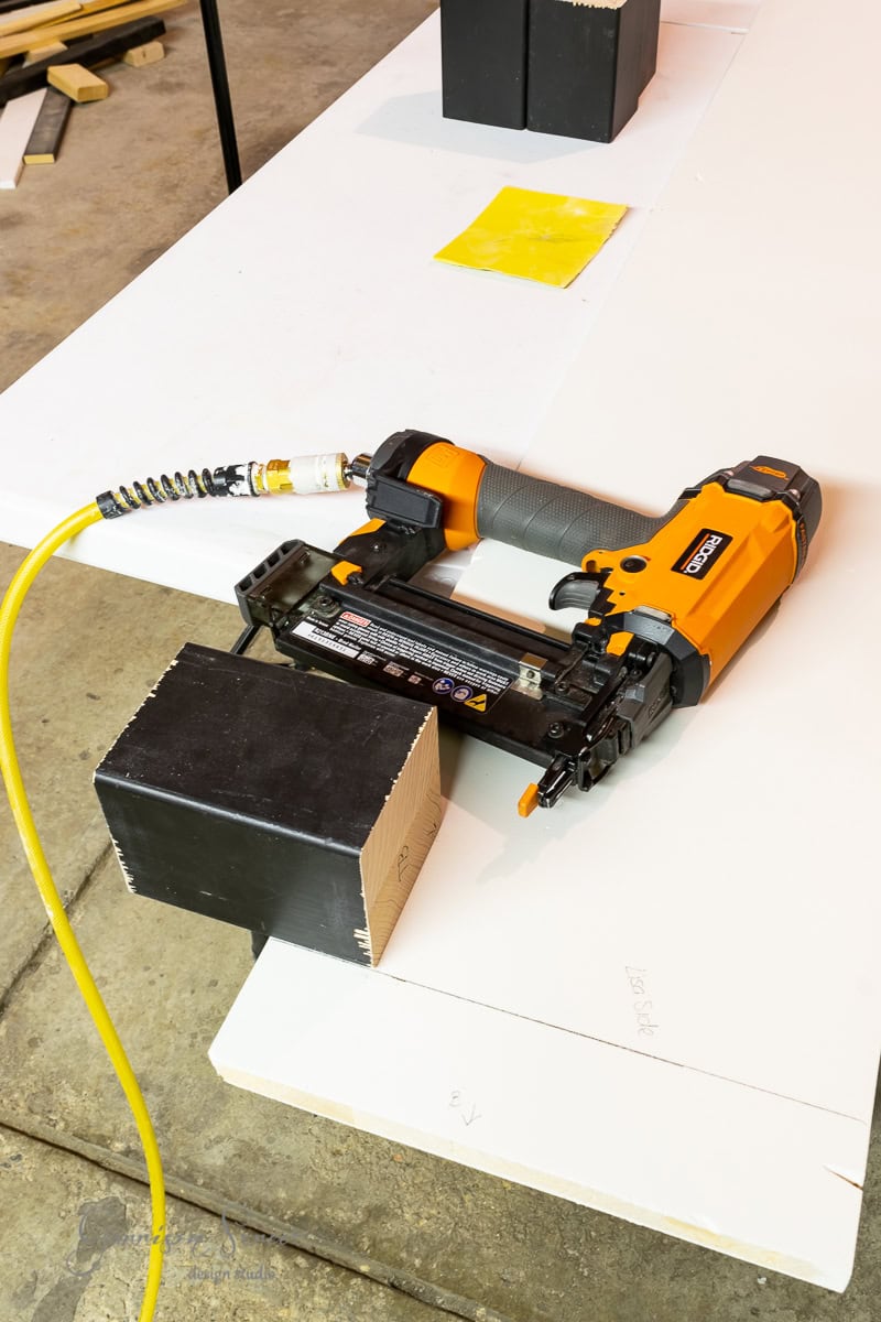 An orange and black pneumatic nail gun rests on a white table, connected to an air hose, ready for assembling projects like a bed frame. A black wooden block and sheets of paper are nearby in the workshop or garage with visible concrete flooring.