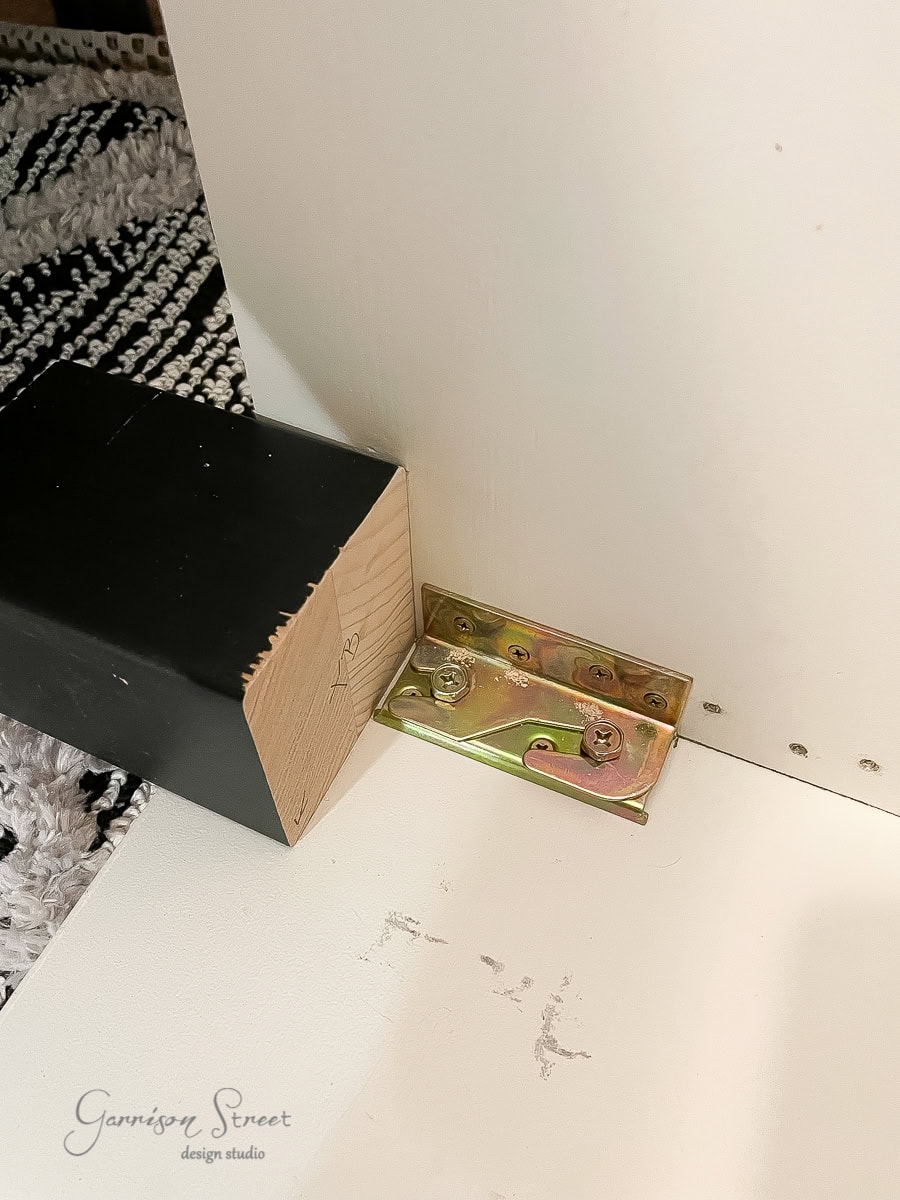 Close-up of a wooden plank from an adjustable base with a black end bracket attached to a wall using a gold metal hinge. The floor features a black and white patterned rug. The wall and floor show minor scuffs and wear. Garrison Street Design Studio logo is visible.