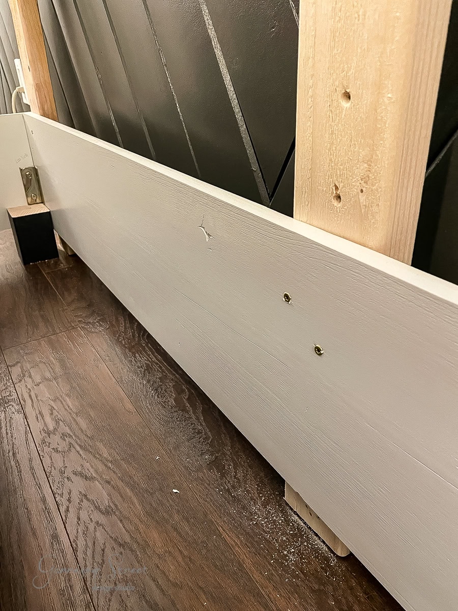 A wooden board is attached to vertical beams on a dark-paneled wall, reminiscent of a sleek bed frame. Two screws are visible on the board. The floor is dark wood, and angled shadows add contrast to the image.