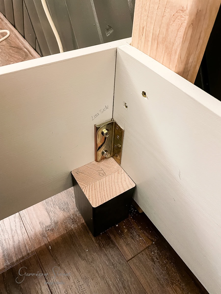 Close-up of a corner construction showing a wooden bed frame leg attached with a metal bracket. The leg is marked "lego side," and the bracket is secured with screws to a white surface, resting on a wooden floor.