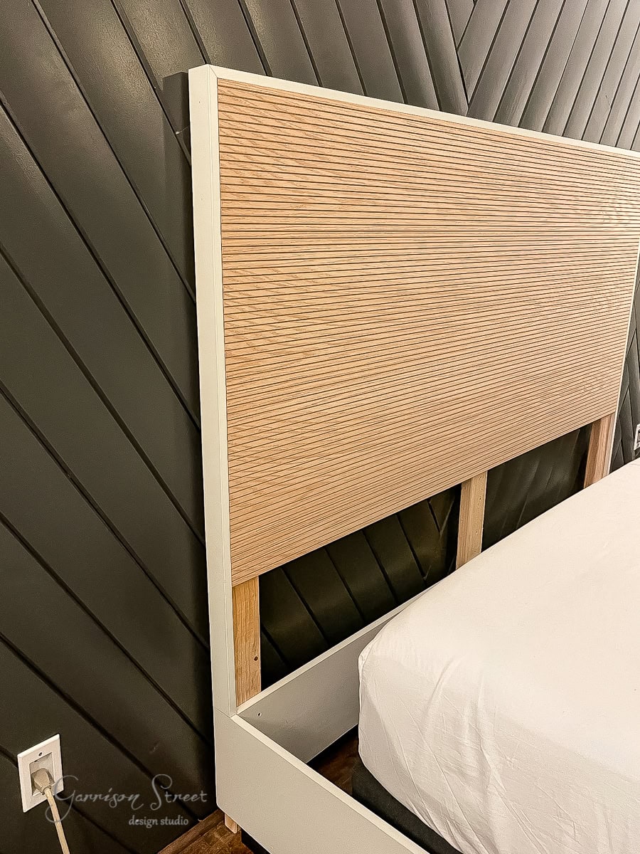 A wooden headboard with vertical slats stands against a dark, diagonally paneled wall. The white bed frame with an adjustable base has a mattress partially visible. An outlet is on the wall, and "Garrison Street Design Studio" is written in the corner.