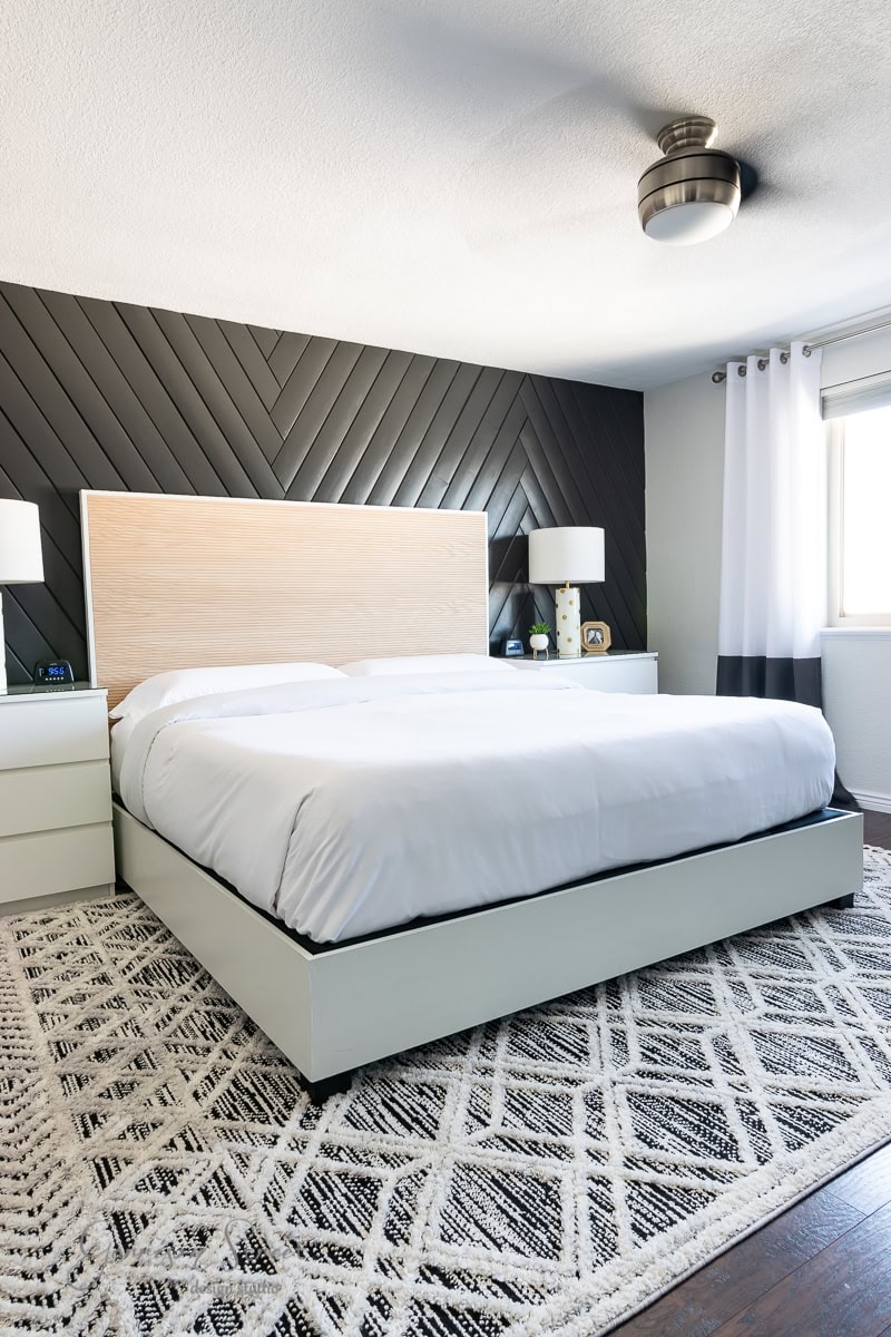 Modern bedroom with a white bed featuring an adjustable base, light wood headboard, and black herringbone accent wall. Two white nightstands with lamps flank the bed. A geometric-patterned rug covers the dark wooden floor. A ceiling fan is above, and a window with white curtains is on the right.