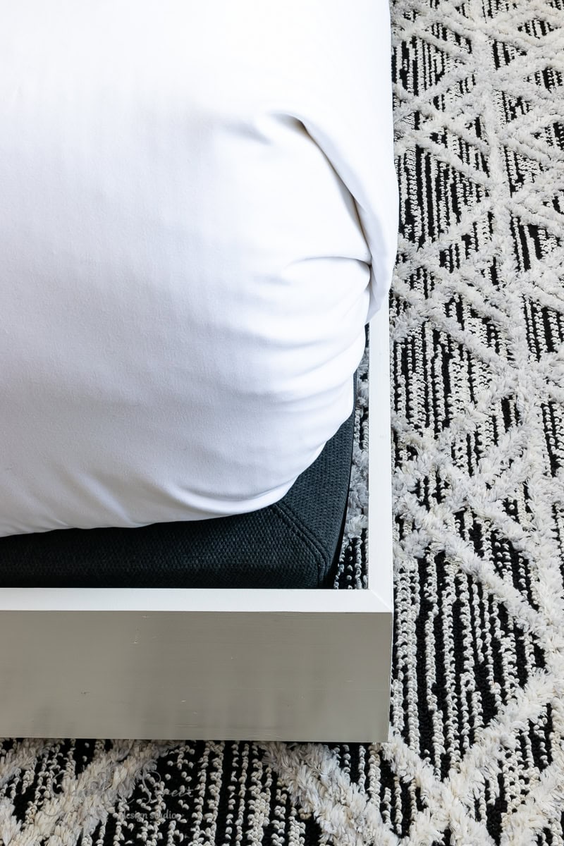 Close-up of a bed corner with white sheets tucked into a light-colored adjustable base. The sleek bed frame rests on a textured black and white patterned rug.