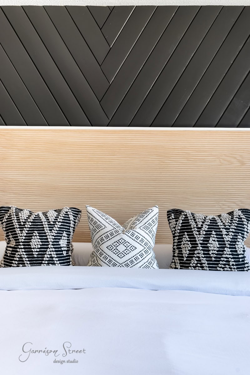 A modern bedroom features a wooden headboard against a black chevron-patterned wall. Three patterned pillows in black and white rest on a neatly made bed with crisp white bedding, atop an elegant bed frame that blends style with functionality.