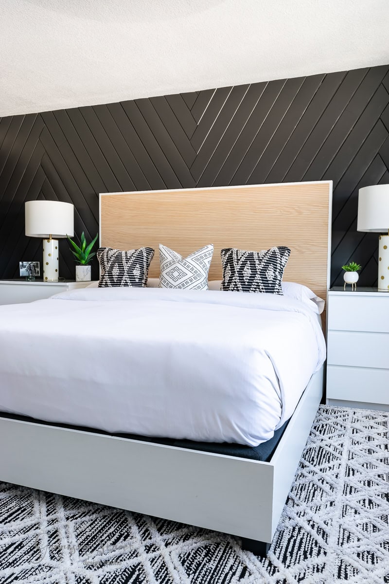 A modern bedroom with a large bed featuring an elegant wooden headboard and white bedding atop an adjustable base. The wall behind showcases a dark, chevron pattern. Two white nightstands with lamps and decorative items flank the bed frame, while a patterned rug adorns the floor.