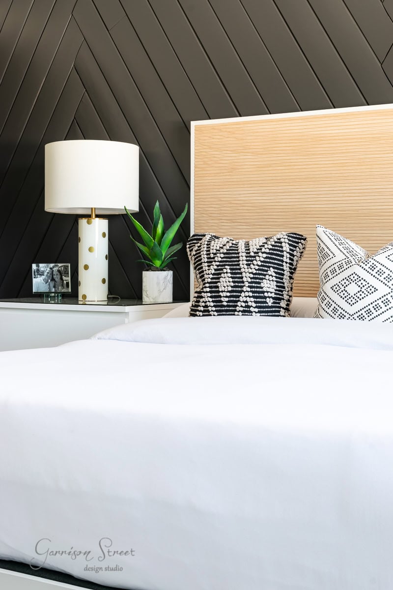 A modern bedroom features a white bed with an adjustable base, complemented by black and white patterned pillows. A nightstand hosts a potted plant and lamp, set against a dark diagonal stripe wall design and light wooden headboard.