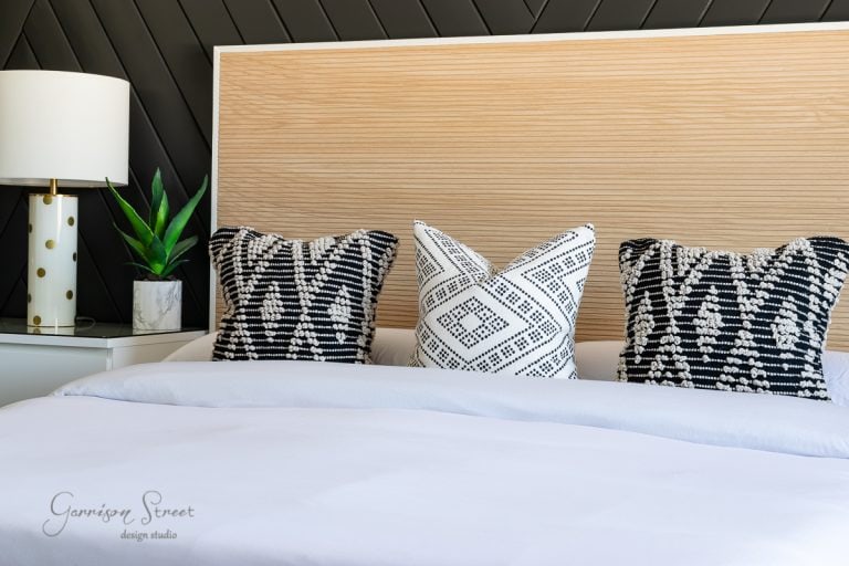 A modern bedroom features a light wooden headboard with an elegant bed frame and three patterned cushions—two black and one white with black designs. A white lamp with a gold base and a small green plant sit on a bedside table. A geometric black wall adds contrast.