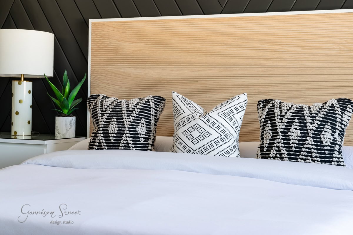 A modern bedroom features a light wooden headboard with an elegant bed frame and three patterned cushions—two black and one white with black designs. A white lamp with a gold base and a small green plant sit on a bedside table. A geometric black wall adds contrast.