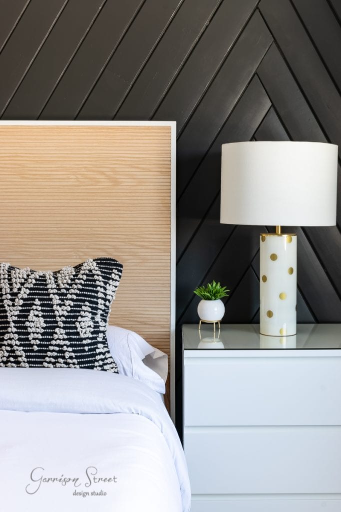 Home A modern bedroom features a wooden headboard set against a black chevron-patterned wall. The white nightstand supports a lamp with gold polka dots and a small plant. A bed frame with an adjustable base complements the white linens and a decorative black-and-white pillow beside it.