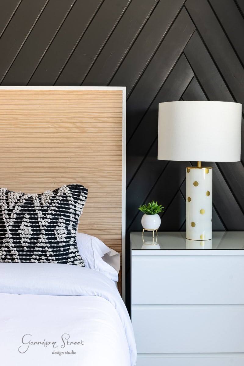 A modern bedroom features a wooden headboard set against a black chevron-patterned wall. The white nightstand supports a lamp with gold polka dots and a small plant. A bed frame with an adjustable base complements the white linens and a decorative black-and-white pillow beside it.