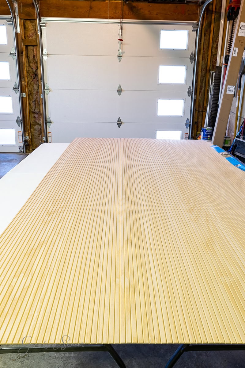 A long, grooved wooden board lies on a table in a garage, possibly part of a bed frame project. The garage door is closed, small windows letting in light. Various tools and materials are visible in the background, suggesting a workshop setting with potential for creating an adjustable base.