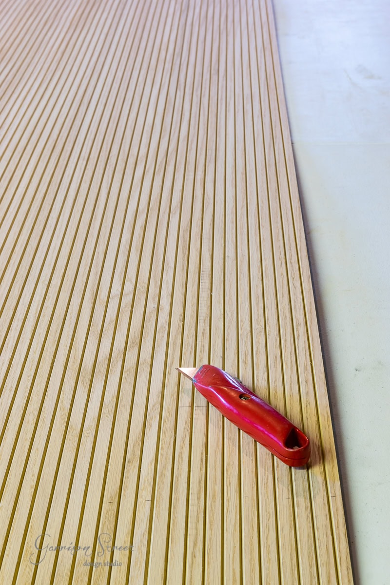 A red utility knife rests on a clean, grooved wooden deck that resembles the precise lines of an adjustable bed frame. The deck occupies most of the frame, with evenly spaced narrow grooves running vertically against a smooth, light-colored background.