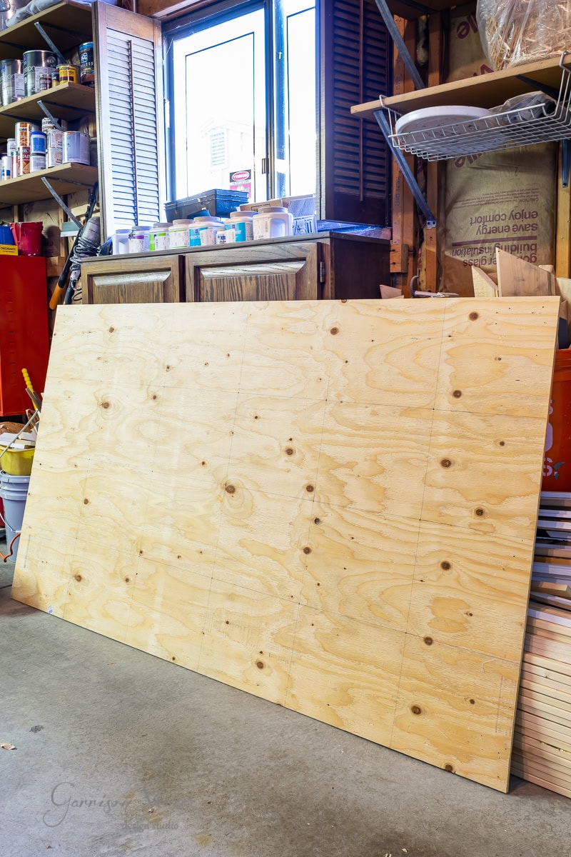 A large sheet of plywood rests on the floor in a workshop, ready to be crafted into a sturdy bed frame. Behind it are shelves with paint cans and tools. Sunlight streams in through a window, illuminating the workspace.