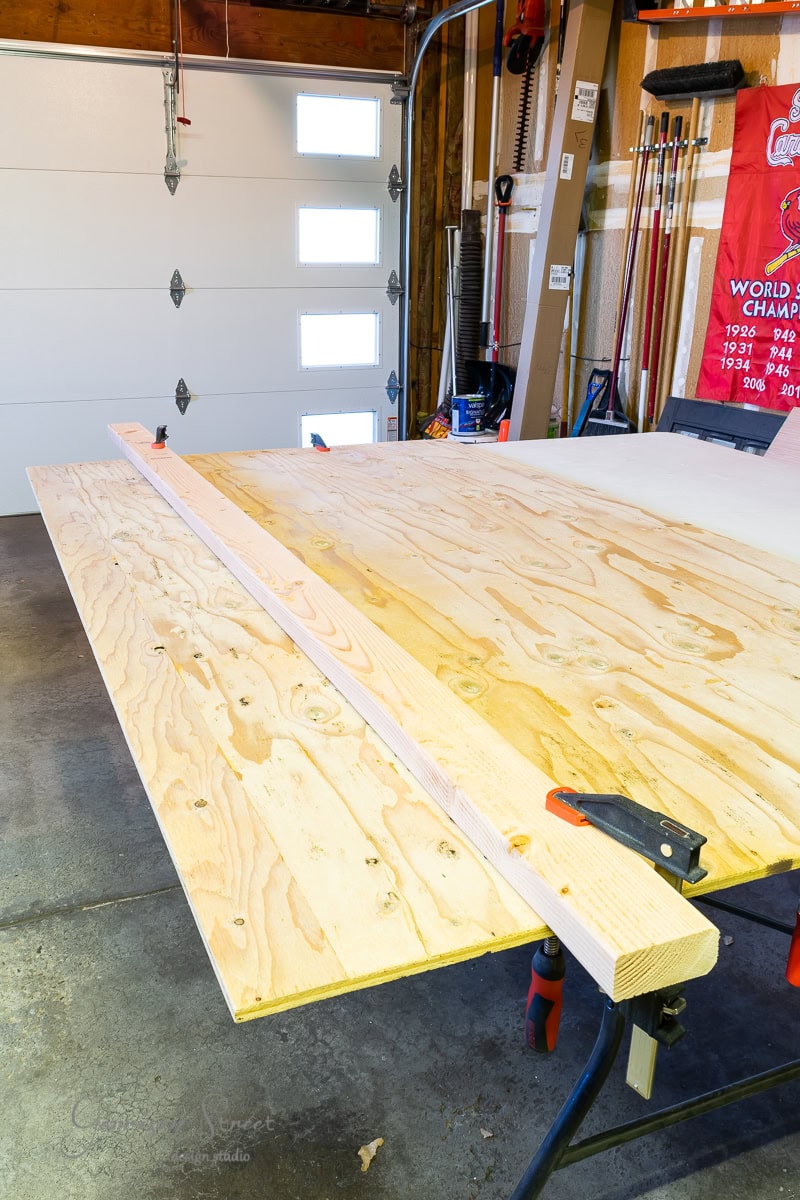 In a woodworking workshop, a large plywood sheet rests on a workbench with a wooden board clamped to it, hinting at the birth of a bed frame. Various tools and equipment are scattered around, alongside an adjustable base and the backdrop of a red banner and garage door.