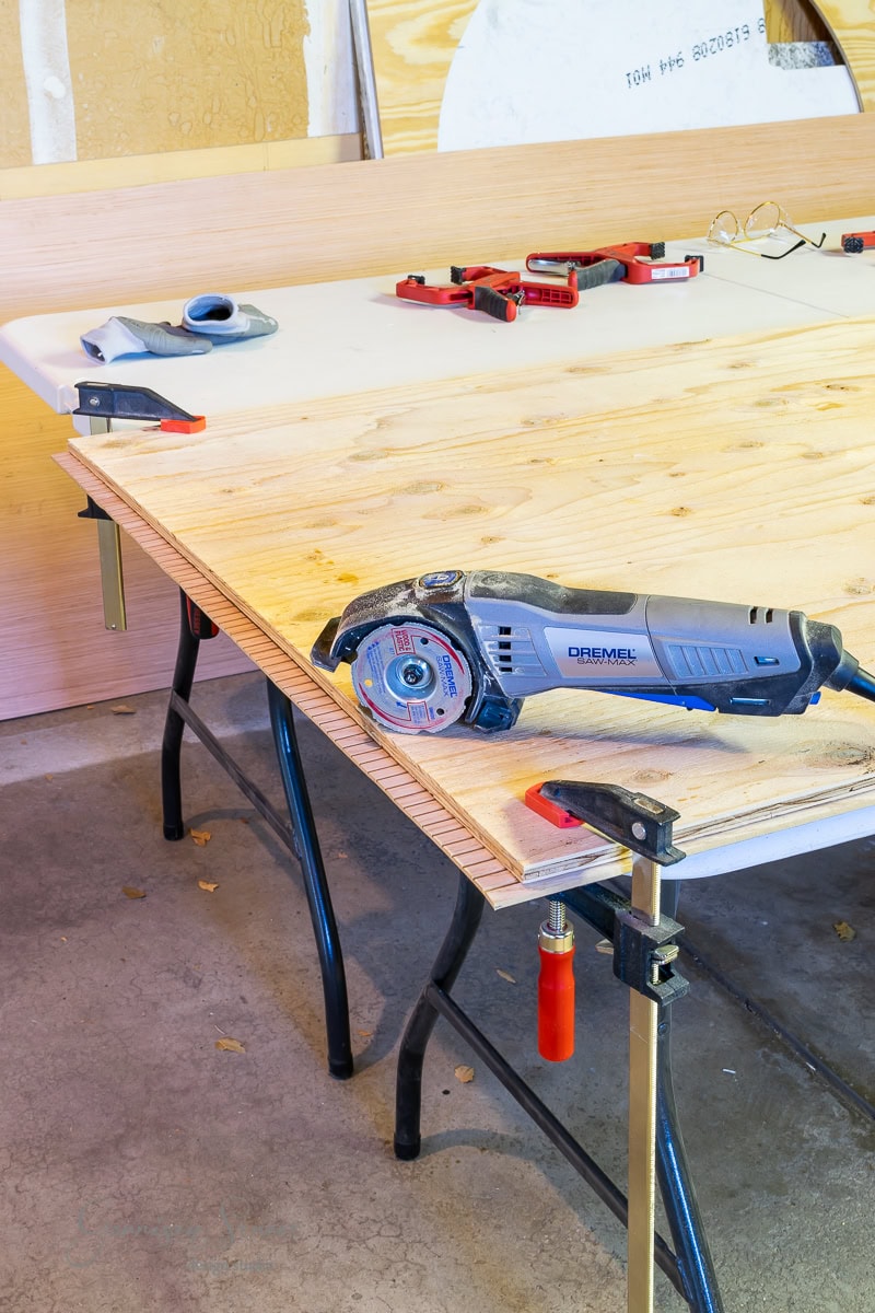 A workbench holds a large wooden board secured with clamps, perfect for crafting a bed frame. On the board is a handheld Dremel saw. Nearby are red tools, safety glasses, and blue gloves. The garage backdrop reveals additional boards and materials, and an adjustable base waiting for assembly.