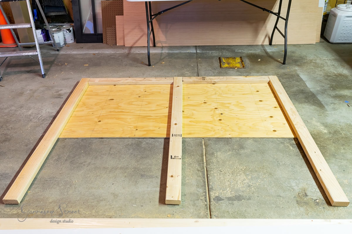 Two wooden frames, soon to become a bed frame with an adjustable base, are laid out on the concrete floor in a workshop. Plywood sheets are attached, ready for the next step. A folding table and various tools are visible in the background.