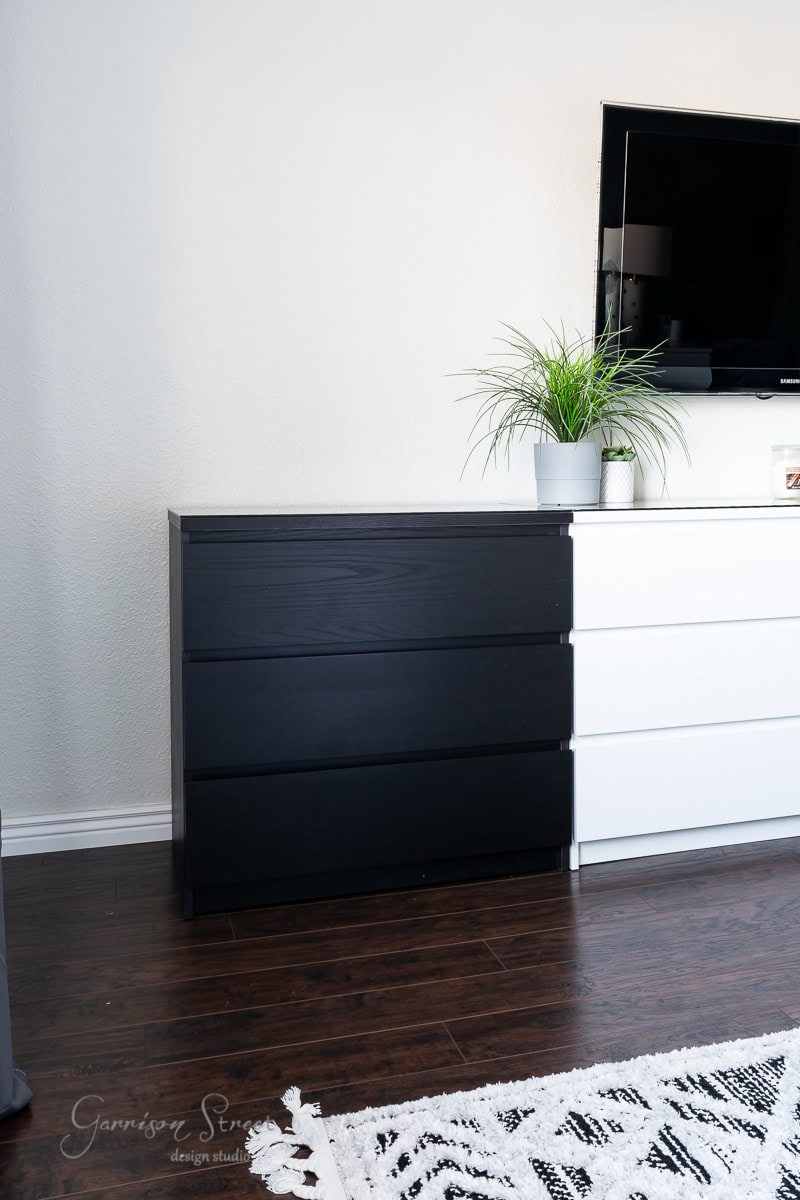 A black IKEA Malm dresser stands beside a white one underneath a wall-mounted TV. A potted plant decorates the white dresser, while a cozy dog crate sits nearby. The room features dark wooden flooring and a patterned rug.