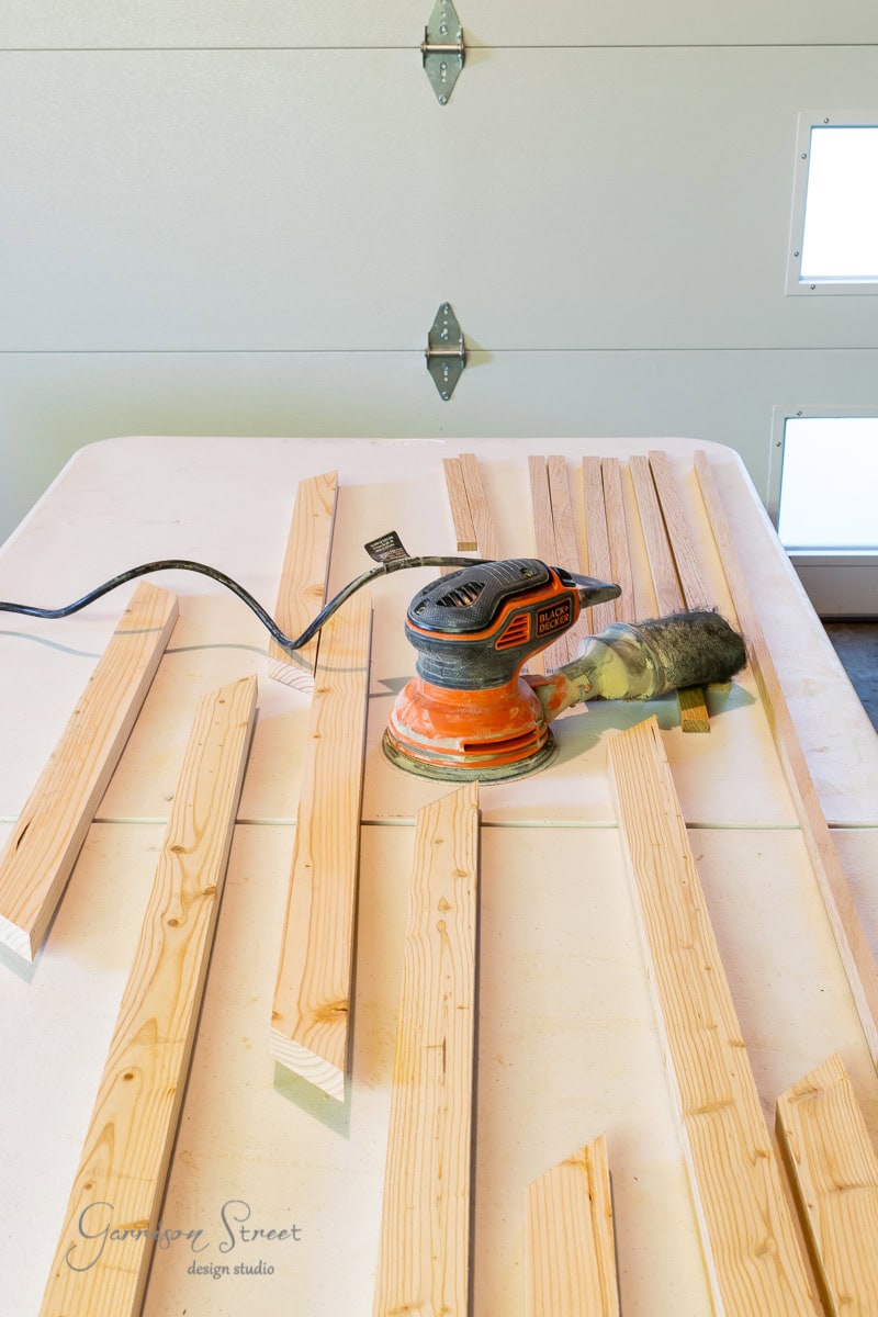 A collection of wooden planks arranged on a table, with an electric sander placed on top. Reminiscent of crafting an IKEA Malm masterpiece, the background features a garage door with a cord extending from the sander. An angled view showcases the wood grain and tool.