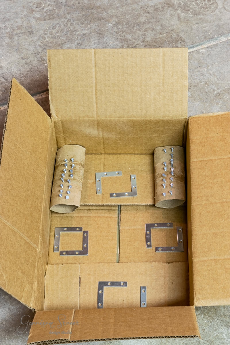 An open cardboard box sits on a stone surface, reminiscent of assembling an IKEA Malm piece. Inside, metal brackets lie flat at the bottom with screws wrapped neatly in paper rolls.