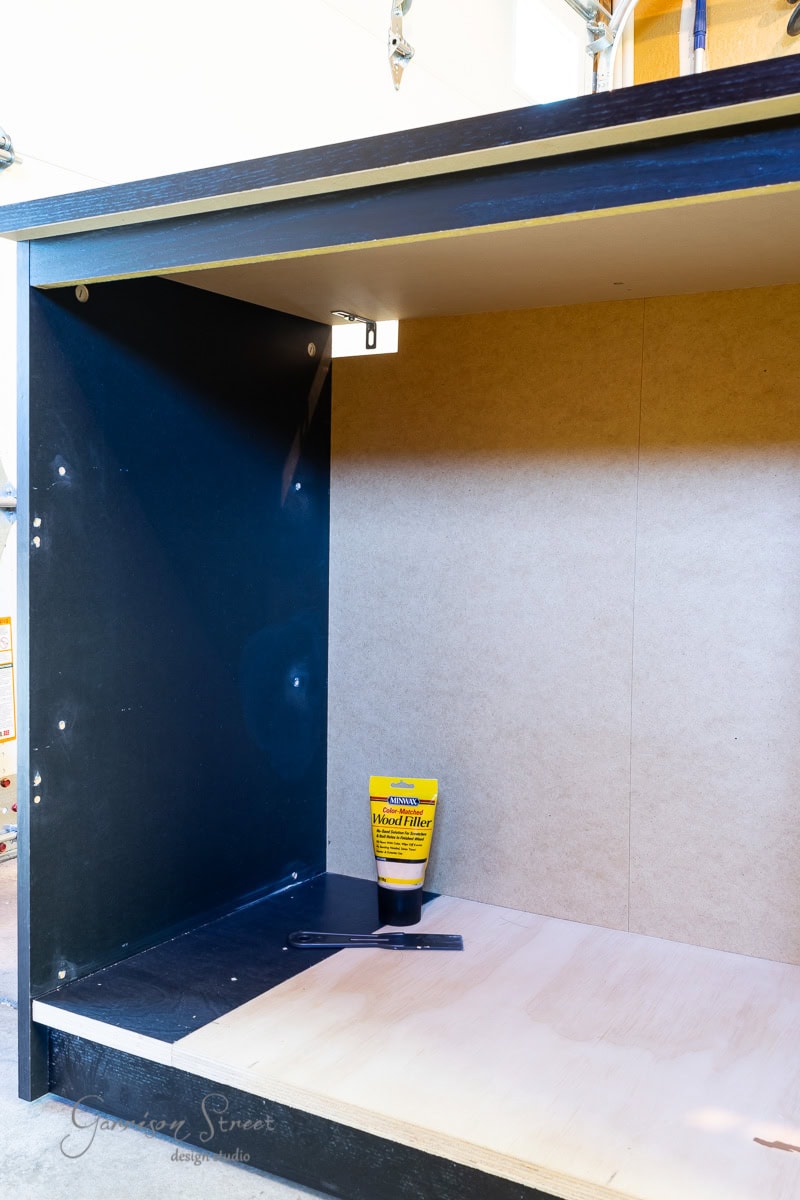 An open wooden cabinet, reminiscent of an IKEA Malm piece, holds a tube of wood filler and a putty knife on its bottom shelf. The cabinet is partially painted black, blending with the garage interior seen in the background.