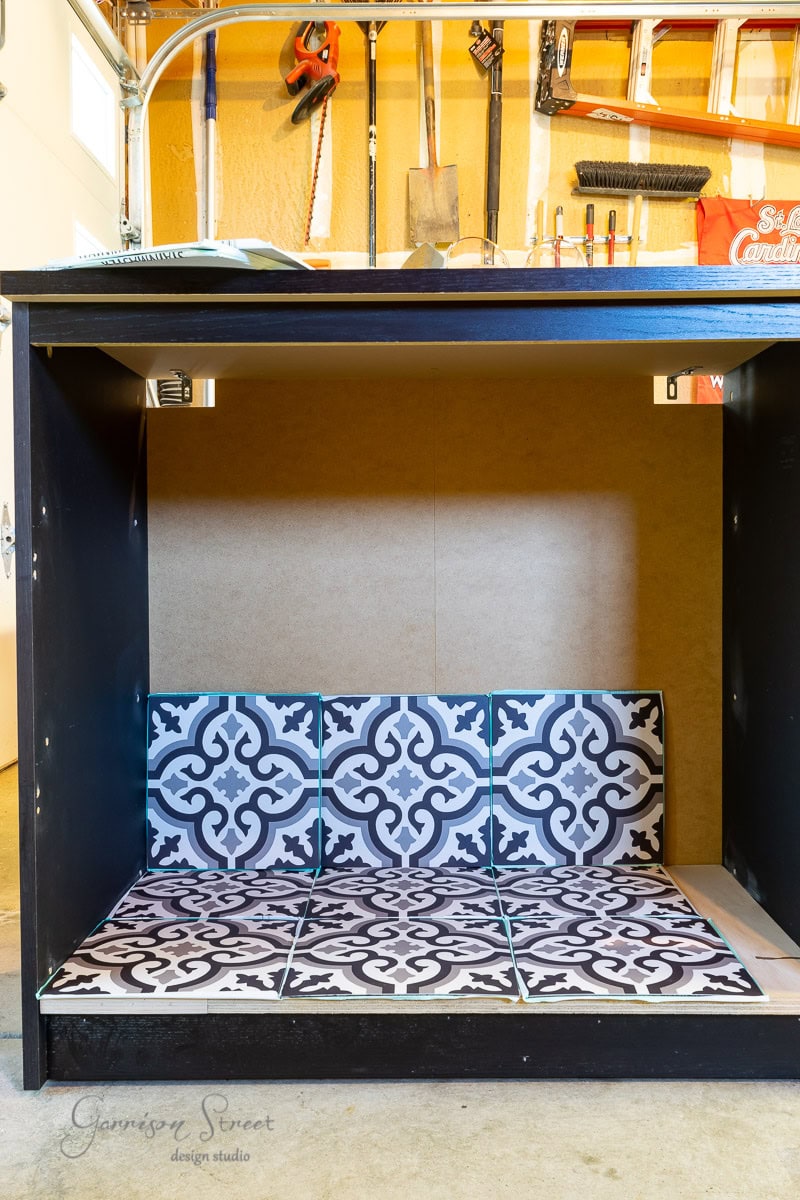 A partially constructed cabinet with a patterned tile backsplash inside, featuring black and white intricate designs, sits atop what looks like an IKEA Malm dresser. Tools and materials are visible in the background, suggesting an ongoing DIY project in a workshop.