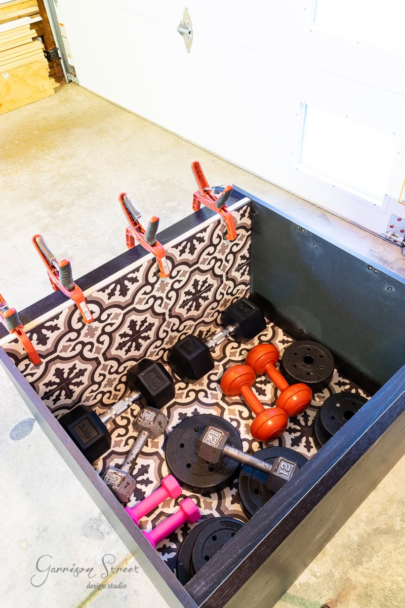 A gym storage box lined with a patterned mat holds various dumbbells, including black and orange weights, much like the efficient design of an IKEA Malm piece. Red clamps are attached to the sides of the box, which sits on a concrete floor in a garage.