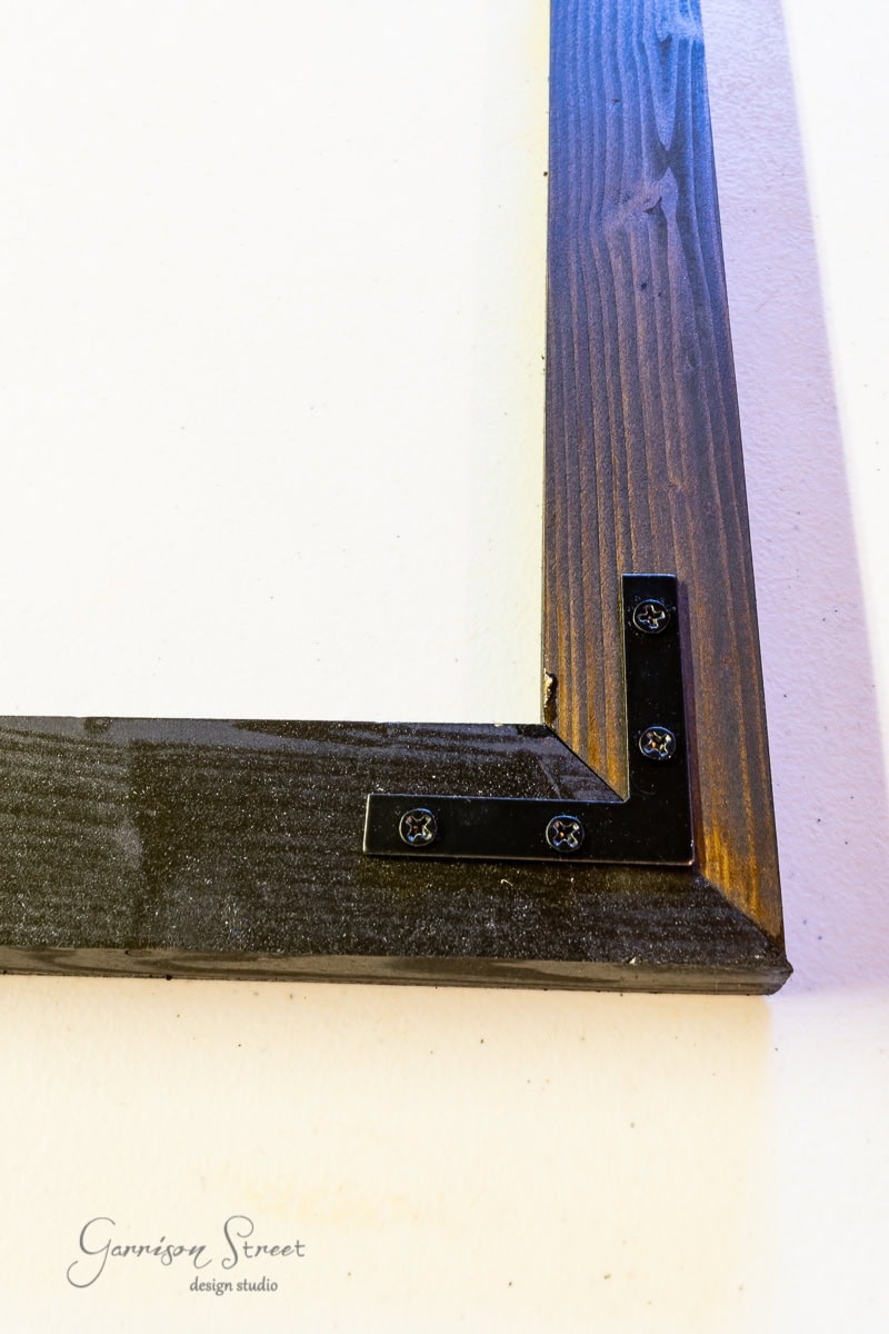 A close-up of a wooden frame corner showcases metal brackets and screws securing the joint. The wood, reminiscent of an IKEA Malm finish, contrasts against a plain white background. At the bottom, "Garrison Street Design Studio" is elegantly inscribed.
