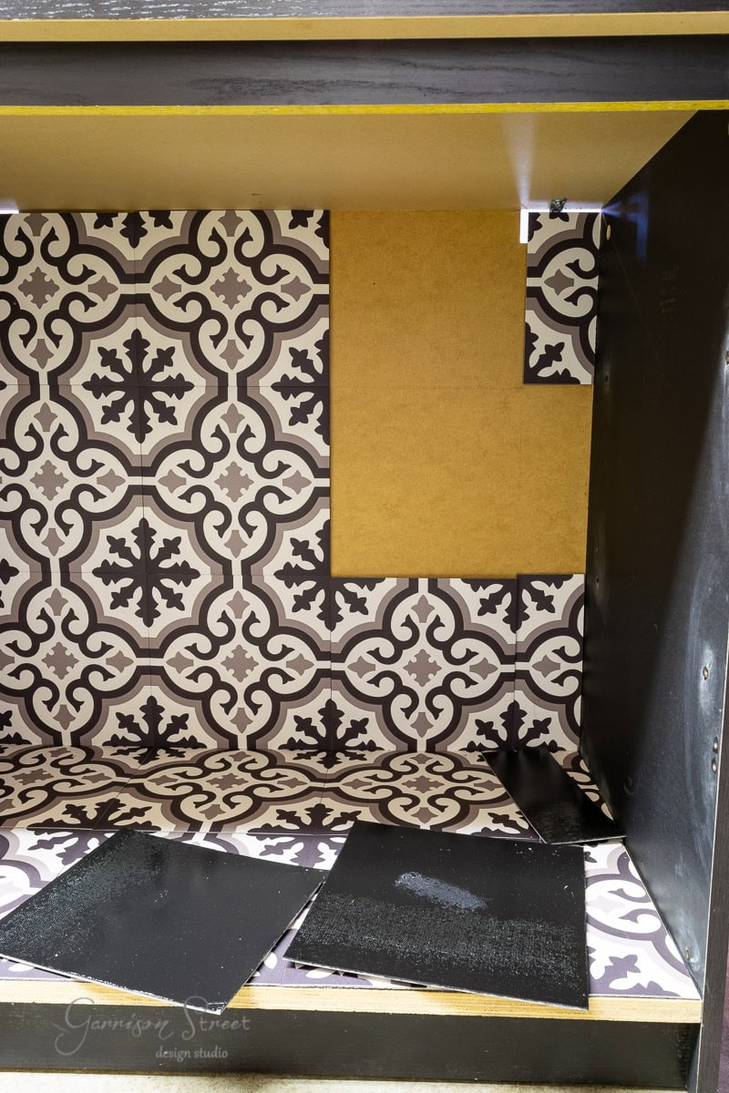 The interior of the cabinet showcases dark wood walls and decorative black-and-white patterned tiles. Some tiles are partially placed, with yellow panels covering parts of the top and side, reminiscent of an IKEA Malm aesthetic. A signature reads "Garrison Street Design Studio.