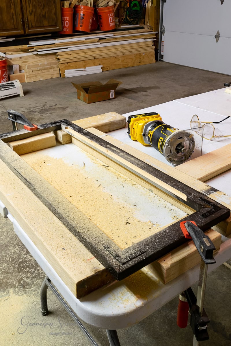 A workshop with woodworking tools on a table, including a yellow power tool, clamps, and a rectangular wooden frame surrounded by sawdust. Stacks of wood are in the background, resembling the precision of an IKEA Malm assembly or crafting a custom dog crate.