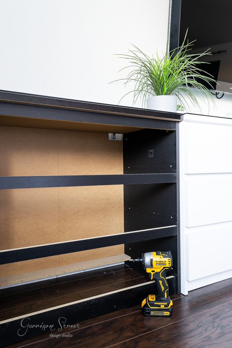A partially assembled black dog crate with an empty frame is shown. A yellow power drill rests on the dark hardwood floor. A white IKEA Malm dresser with a potted plant on top is partially visible on the right.