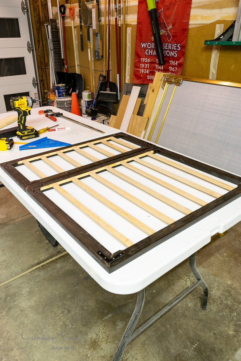 A wooden frame resembling an IKEA Malm piece with horizontal slats rests on a table in a garage workshop. Tools and materials, including a drill and wood pieces, are scattered about. A red "World Series Champions" banner proudly hangs in the background.