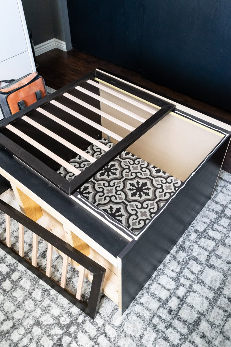 A partially assembled IKEA Malm wooden bed frame features a decorative black and white patterned panel. Positioned on a gray patterned rug, it shares the space with an orange bag of tools nearby, resembling the organized charm of a sleek dog crate setup.