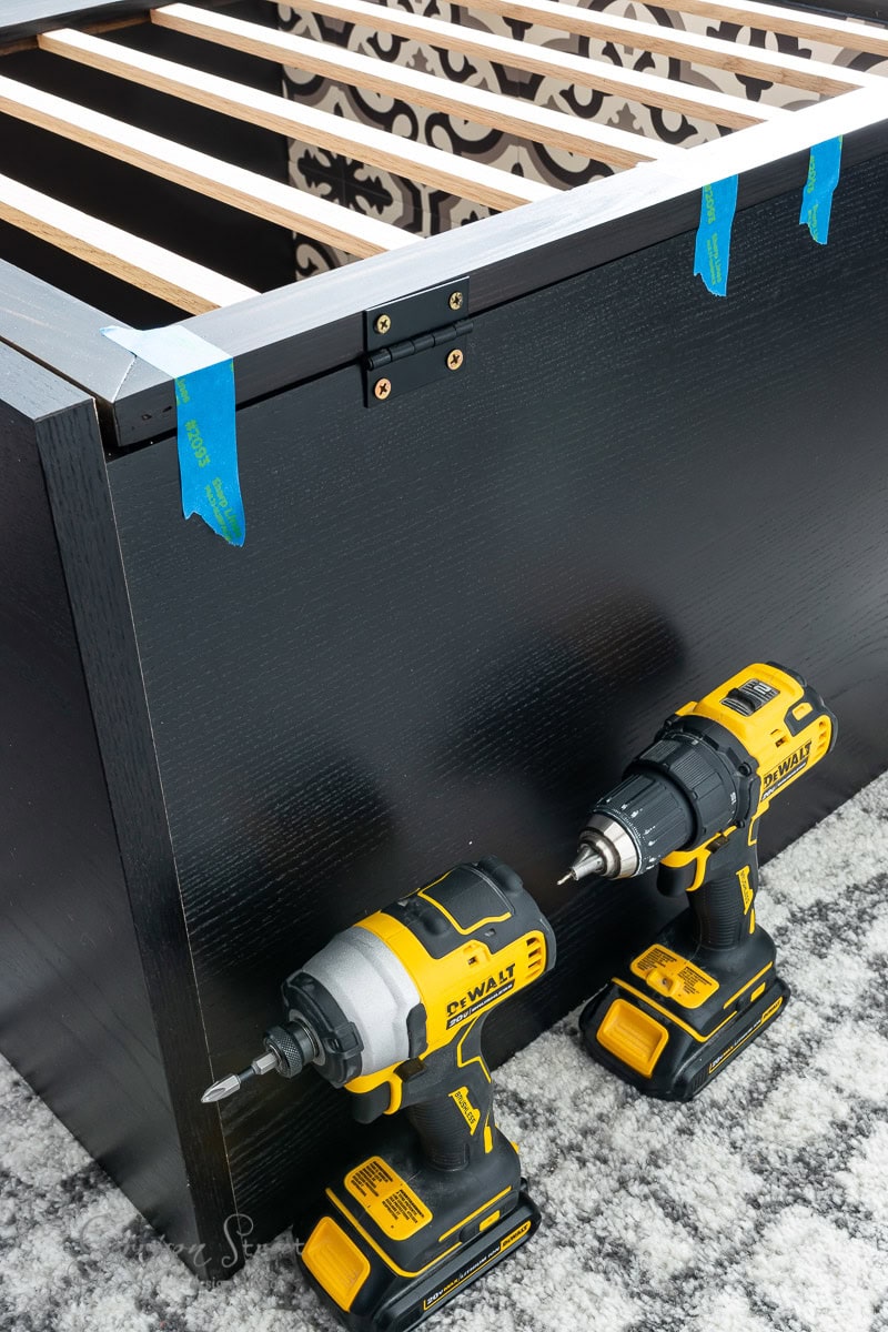 Two yellow and black power drills rest against a partially assembled black bed frame, reminiscent of an IKEA Malm. Blue painter's tape marks positions on the frame. The background reveals patterned wallpaper and a carpeted floor, creating a cozy setting possibly near a dog crate.
