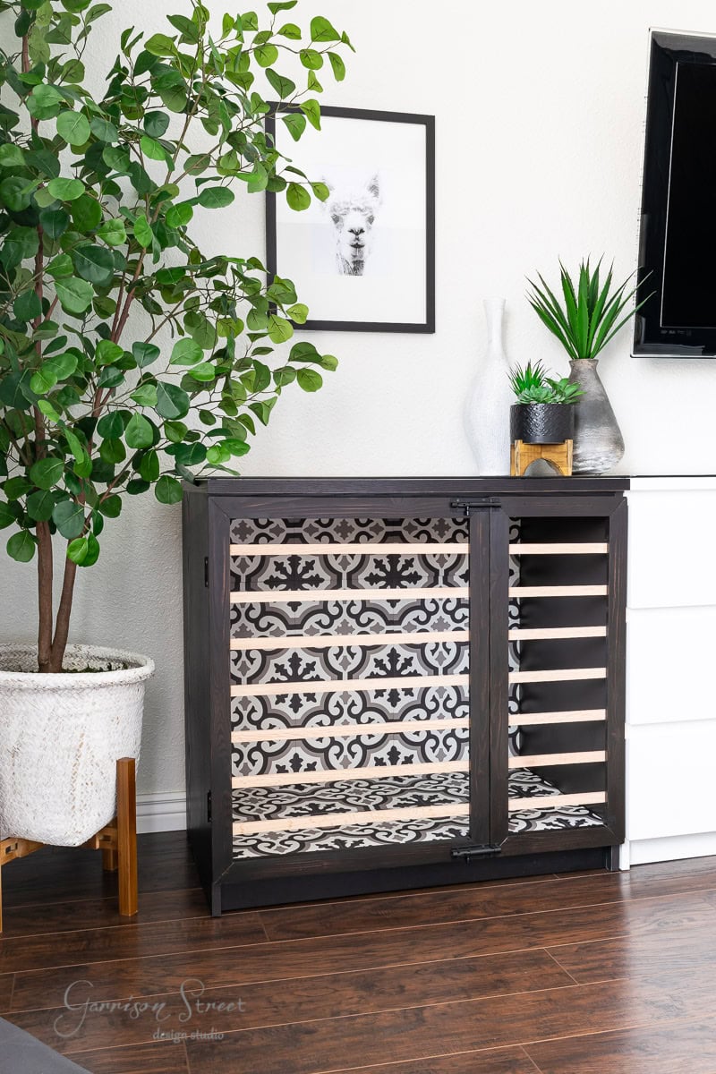 A stylish black wine rack with a decorative patterned back and light wood shelves stands in a living room, complementing an IKEA Malm dresser. Beside it is a potted tree and a white cabinet. Above the rack hangs a framed picture of a llama, with a TV partially visible on the right.