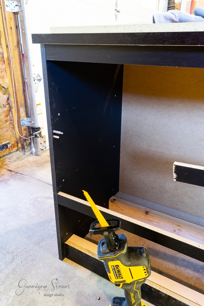 A yellow power saw rests on a partially constructed black wooden cabinet, reminiscent of an IKEA Malm design, in a workshop. The cabinet is open at the front, showing unfinished shelving. Tools and woodwork materials are visible in the background, indicating ongoing construction.