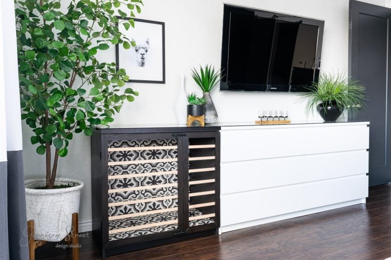 Modern living room corner featuring a black wine rack with patterned shelves and a large leafy plant beside it. An IKEA Malm dresser with a flat-screen TV and potted plants sits against a light wall adorned with minimalist art. A cozy dog crate blends seamlessly into the room's dark wood floor.