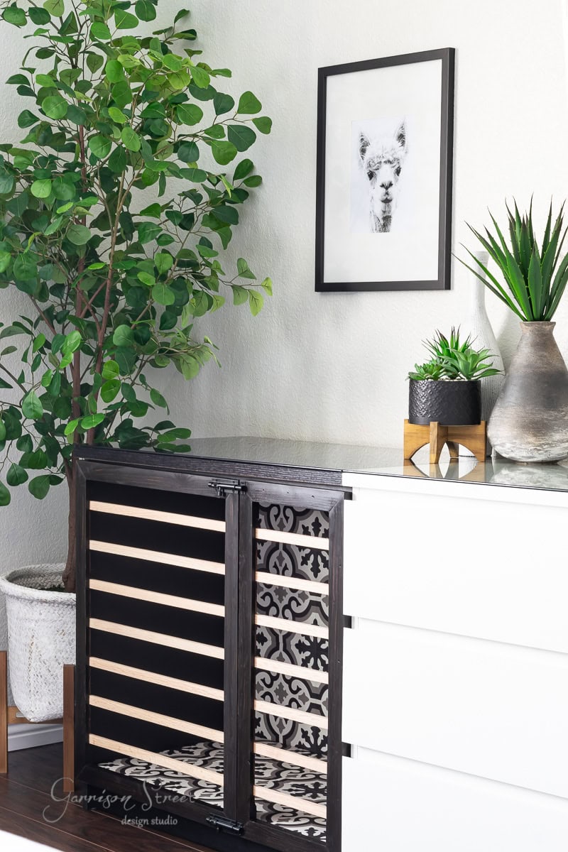 A modern room features an IKEA Malm dresser alongside a wooden wine rack with horizontal shelves. A framed llama portrait hangs above, while potted plants like a small tree and succulents add greenery. Nearby, a stylish dog crate seamlessly blends into the decor.