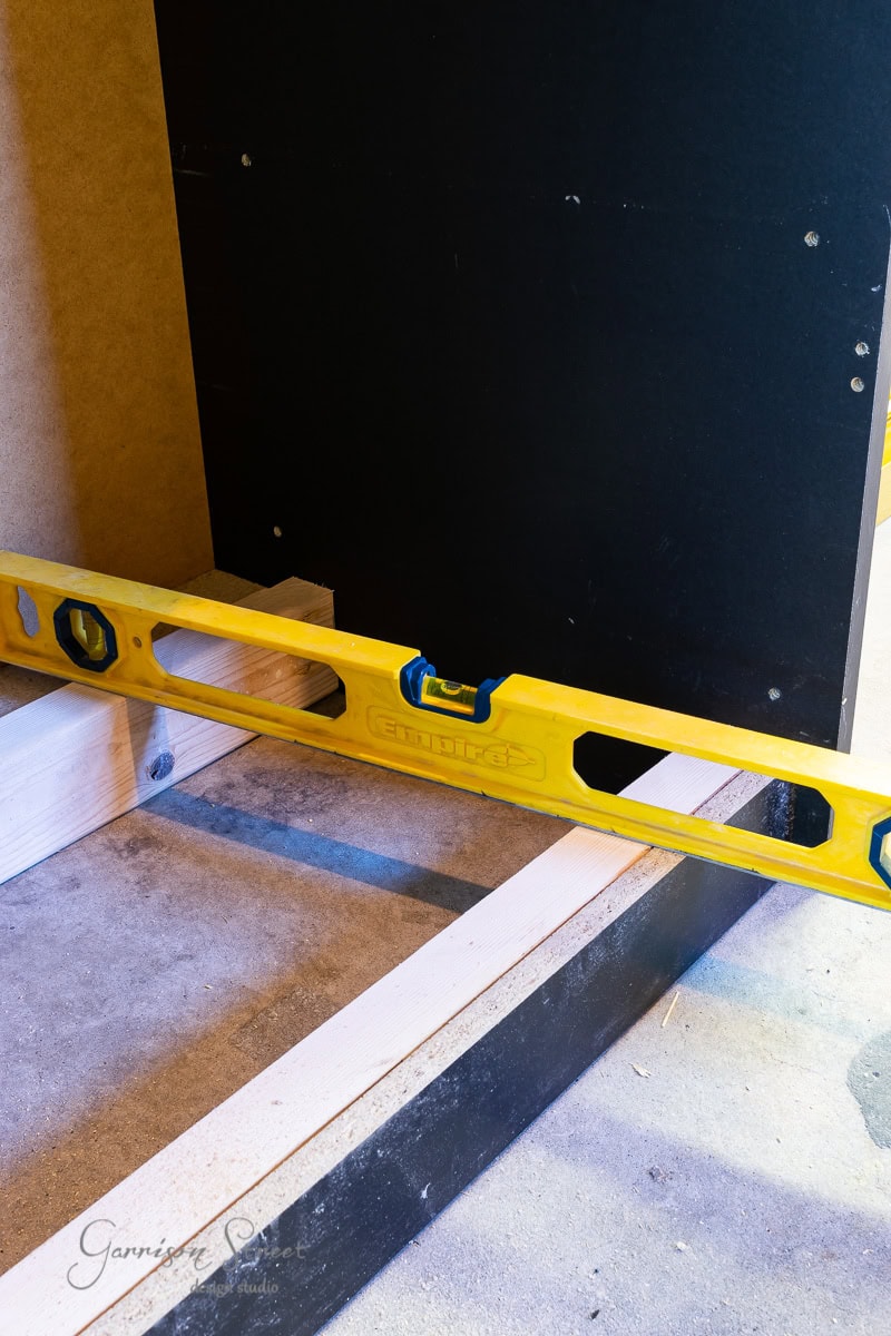 A yellow spirit level rests on a wooden beam, perfectly aligned with another against a black vertical surface in the workshop. Nearby, markings are visible on the concrete floor, reminiscent of an assembly space for an IKEA Malm or even constructing a custom dog crate.