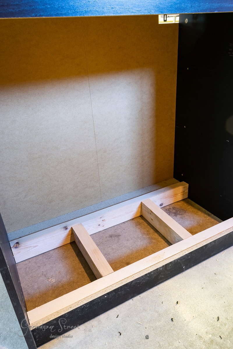 An unfinished wooden cabinet interior with bare wood supports at the bottom resembles the minimalist design of an IKEA Malm piece. The plain back panel reveals the structure behind potential doors, while wood shavings scattered on the floor suggest it could double as a cozy dog crate.