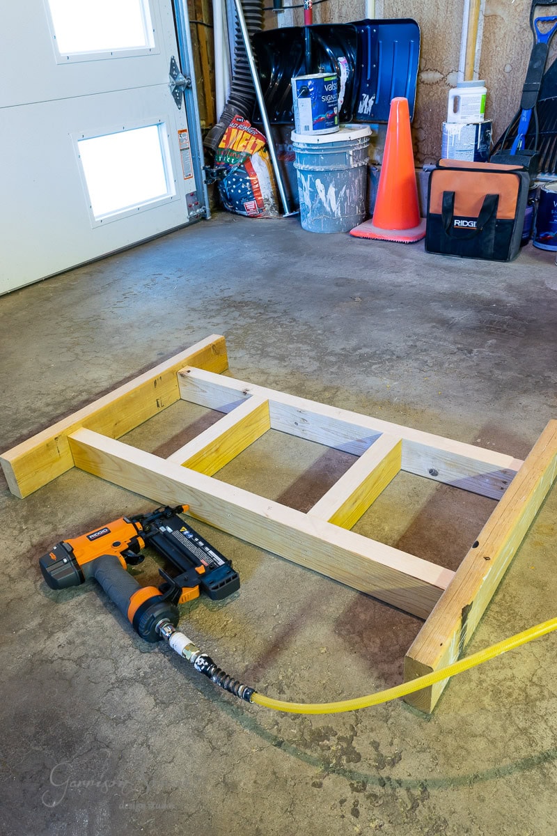 A wooden step ladder frame lies on a workshop floor. Nearby, an orange pneumatic nail gun connects to a yellow hose. Various tools and items are visible in the background, including an IKEA Malm piece and a toolbox, all under the watchful eye of a traffic cone.