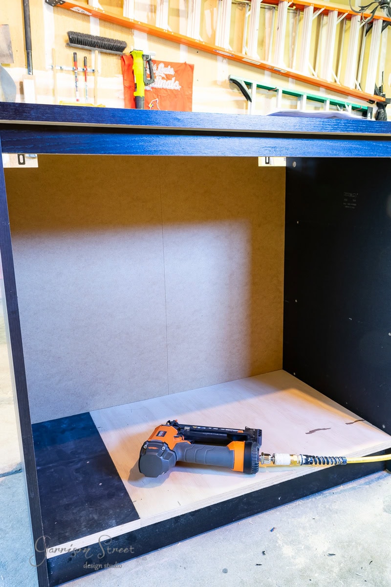 A partially assembled cabinet resembling an IKEA Malm, with a wooden interior and black exterior, stands in a workshop. An orange and black nail gun connected to an air hose rests inside like it's waiting to build a cozy dog crate. Tools and shelves are visible in the background.