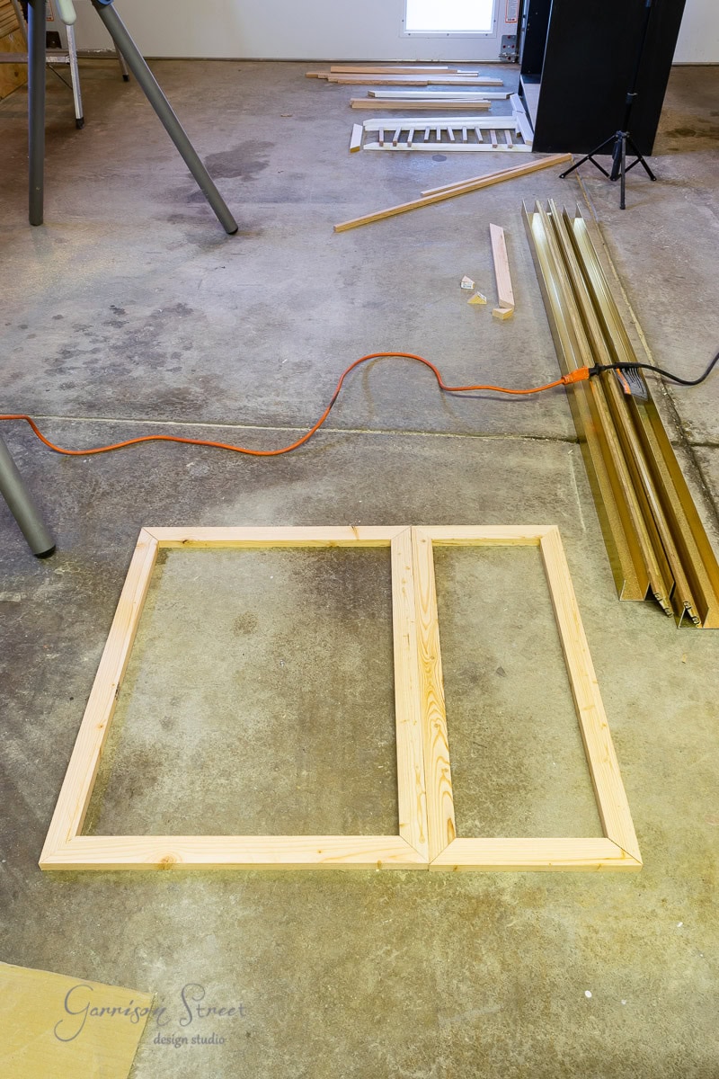 A workshop floor with wooden frames laid out, surrounded by wooden planks and an orange extension cord, resembles an assembly zone for a custom dog crate. A small window is visible in the background, casting light on what could easily be DIY IKEA Malm assembly pieces.