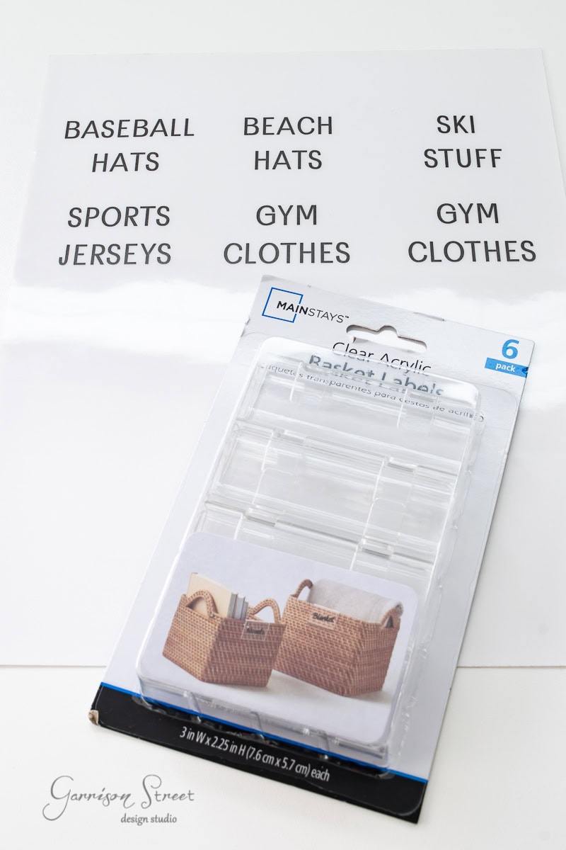 A pack of six clear acrylic basket labels lies on a surface beneath a sheet with black text labels for organizing your reach-in closet or IKEA Pax: "Baseball Hats," "Beach Hats," "Ski Stuff," "Sports Jerseys," "Gym Clothes," and "Gym Stuff.
