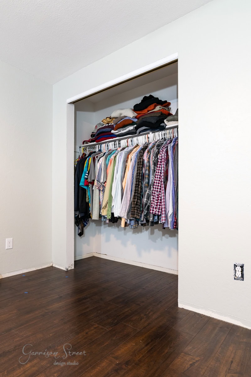 The open reach-in closet boasts a simple design with a light-colored interior and dark wooden flooring. A variety of shirts and jackets hang on the rod, while shelves above house neatly folded clothes, reminiscent of an IKEA Pax setup.