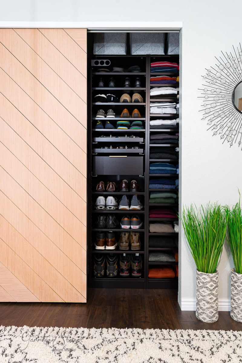 A neatly organized reach-in closet with wooden sliding doors showcases shoes on multiple shelves and folded clothes stacked above. Decorative plants and a round mirror with a sunburst frame adorn the adjacent wall, making it feel like a stylish IKEA Pax design.