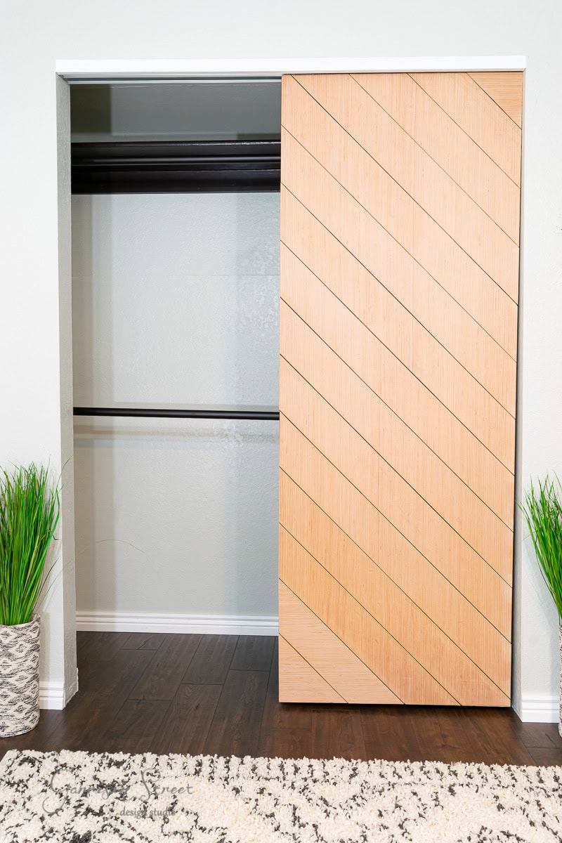 A modern reach-in closet with a partially open wooden sliding door boasting diagonal lines. Inside, two horizontal black metal rods offer space. The floor is dark wood, complemented by green potted plants on either side of the opening, reminiscent of an IKEA Pax setup.