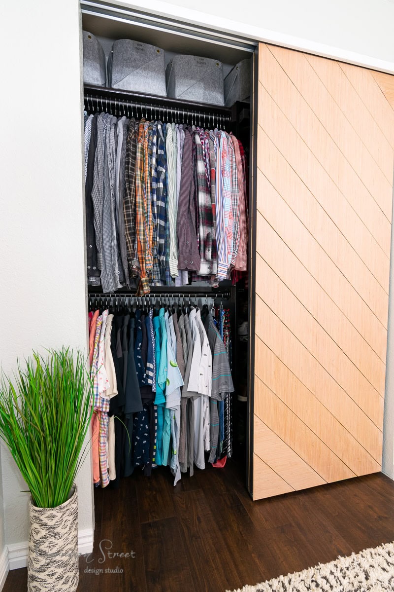 A neatly organized IKEA Pax reach-in closet features two rows of hanging clothes, including shirts in various colors and patterns. Several gray storage boxes reside on the upper shelf, while a decorative plant stands next to the closet with a stylish sliding door.