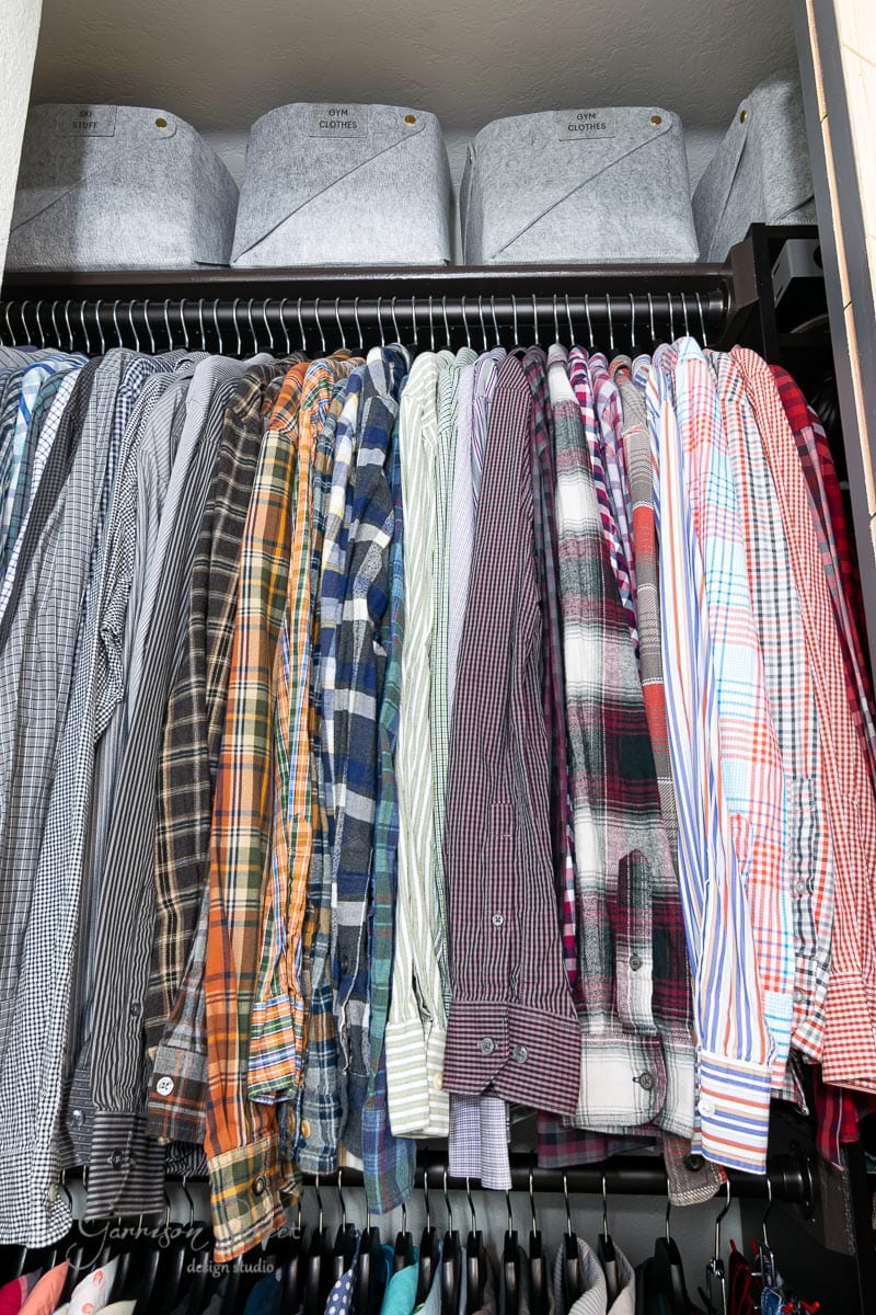 A reach-in closet features a row of neatly hung plaid and striped shirts. Above the shirts, four gray IKEA Pax storage boxes labeled "Extra Blankets" and "Christmas Decor" are visible.